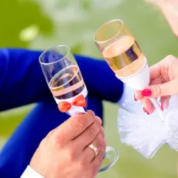 Dwie osoby trzymają kieliszki szampana udekorowane kokardką i tiulem, w tle zieleń, widoczny pierścionek na palcu mężczyzny.