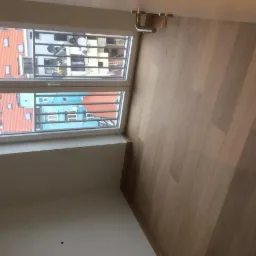 Jasne panele podłogowe ułożone wzdłuż okna balkonowego z widokiem na budynki mieszkalne z czerwonymi i niebieskimi dachami.