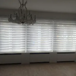 Wnętrze pokoju z trzema oknami przysłoniętymi białymi, poziomymi roletami typu dzień i noc, pod oknami grzejniki, na suficie ozdobny, kryształowy żyrandol