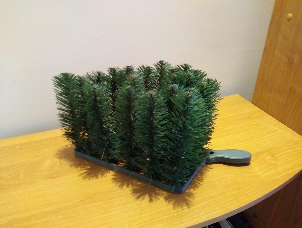 Sztuczne choinki iglaste w plastikowej, szarej podstawce z uchwytem, ustawione na drewnianym blacie.