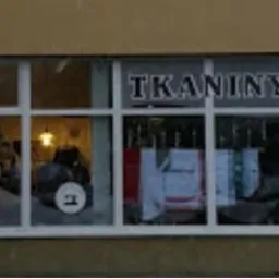 Fasada sklepu z tkaninami i usługami krawieckimi, widoczne napisy 'Tkaniny', 'Usługi krawieckie', 'Szycie na miarę', 'Poprawki', oraz logo z igłą i nitką.