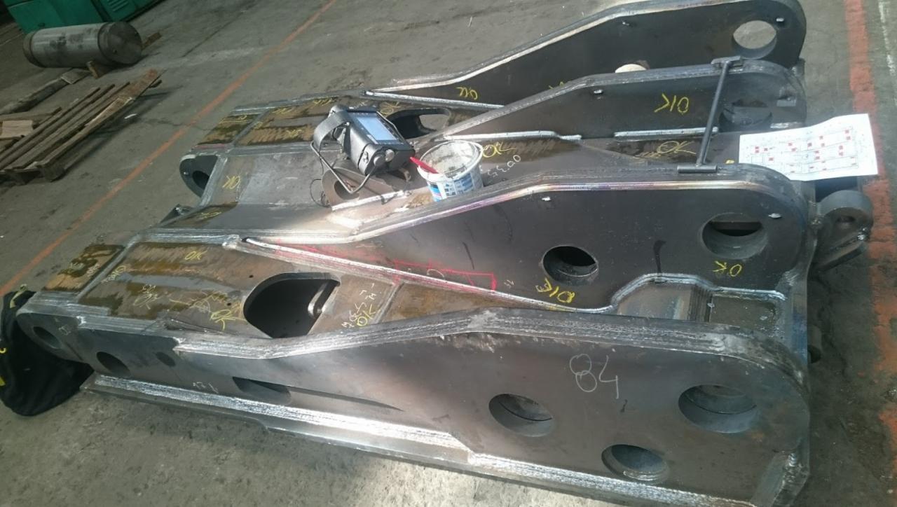 Części konstrukcji stalowej z widocznymi spawami, leżące na podłodze hali, obok leży urządzenie pomiarowe i puszka farby.