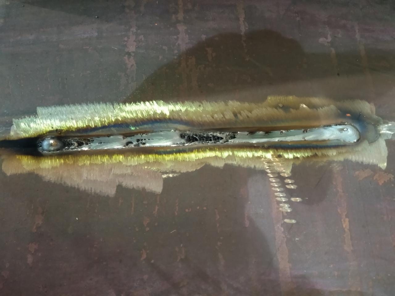 Spoina spawalnicza na ciemnej blasze stalowej, widoczne nierówności i przebarwienia termiczne.