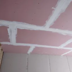 Różowe płyty gipsowo-kartonowe na suficie z widocznymi spoinami wypełnionymi białą masą szpachlową, widoczne drewniane elementy konstrukcji.