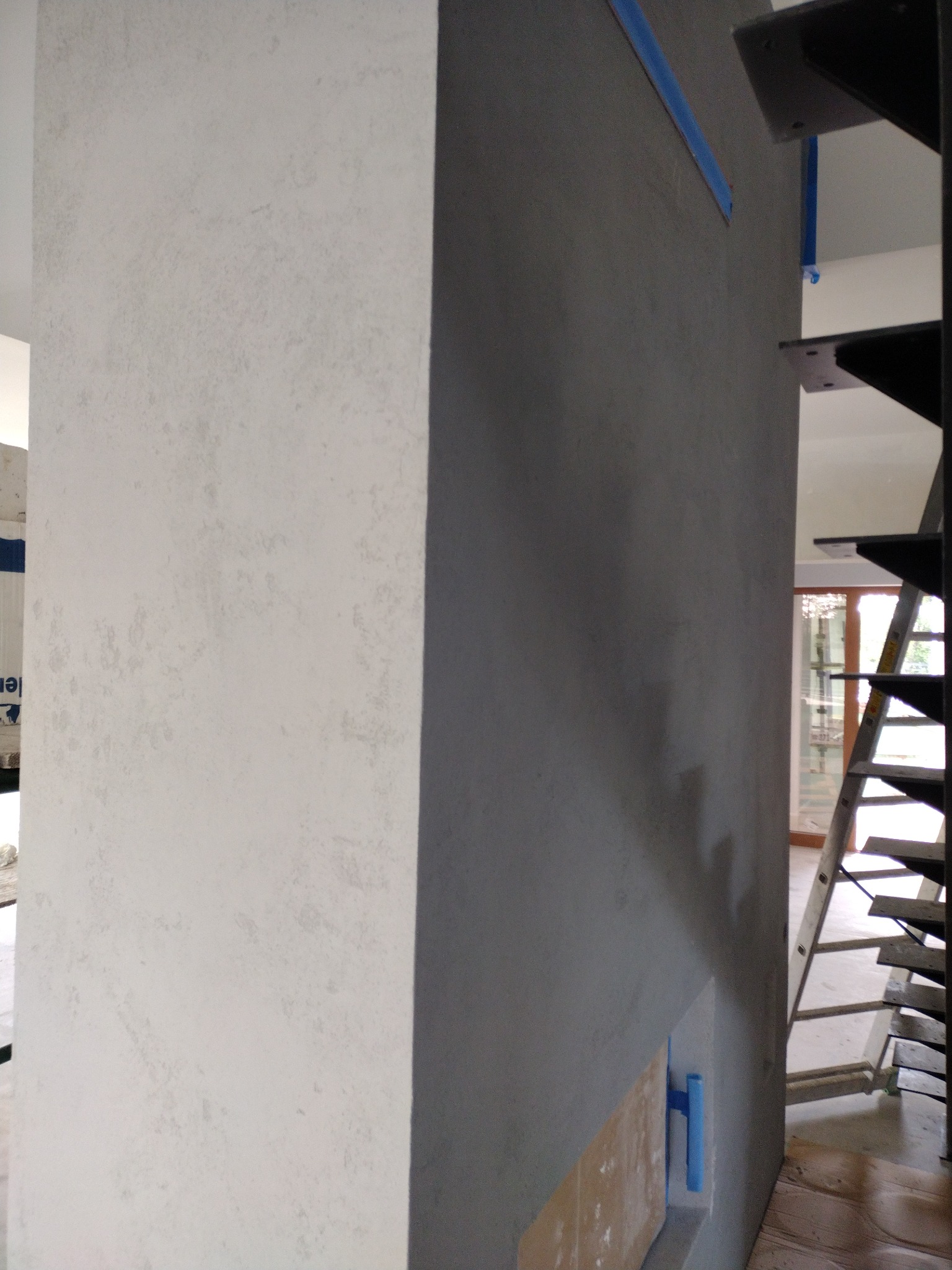 Ściana z efektem betonu ozdobnego w trakcie wykańczania, widoczna szpachelka z niebieskim uchwytem, schody i drabina w tle.