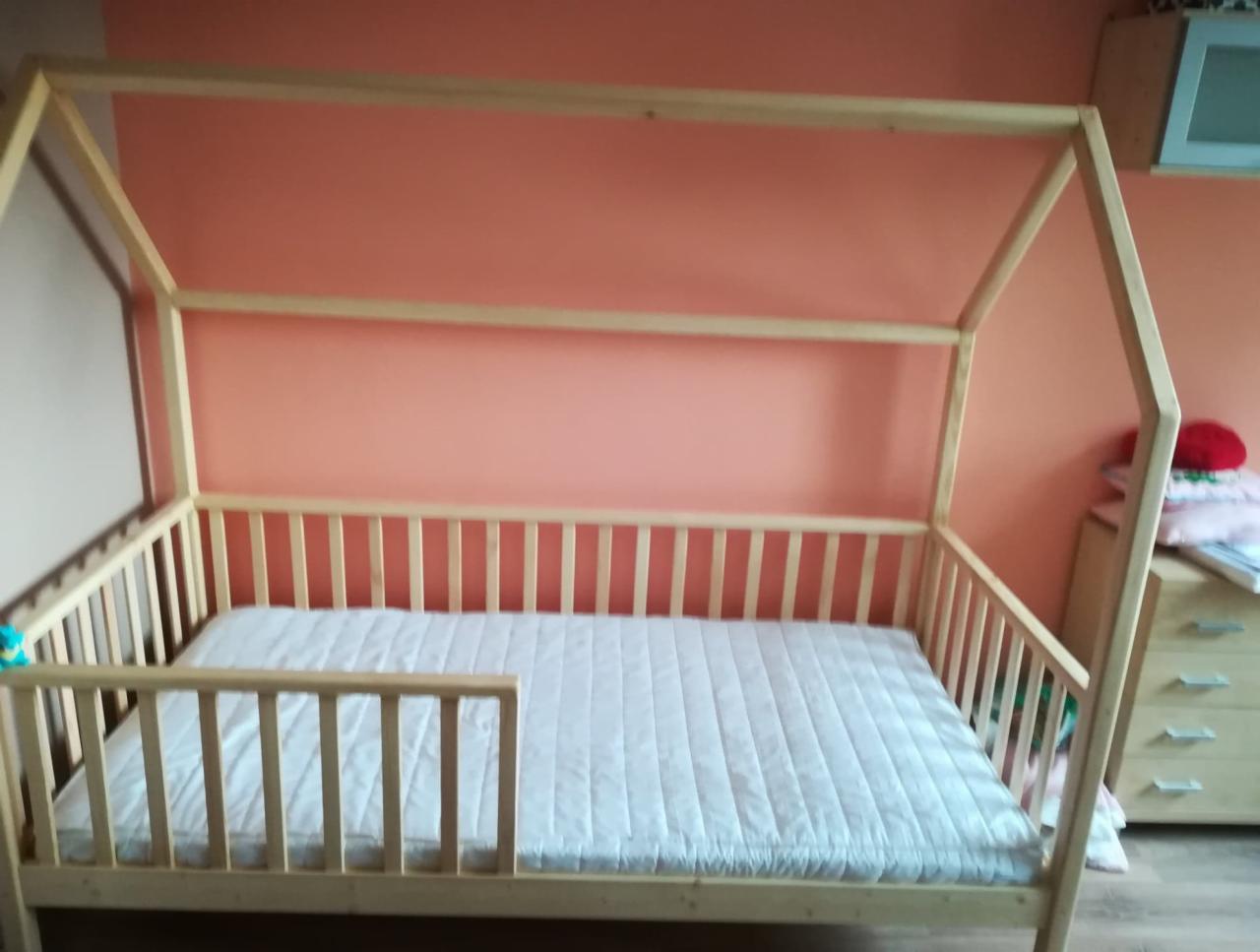 Drewniane łóżko dla dziecka w kształcie domku, z barierką zabezpieczającą i materacem, na tle pomarańczowej ściany.