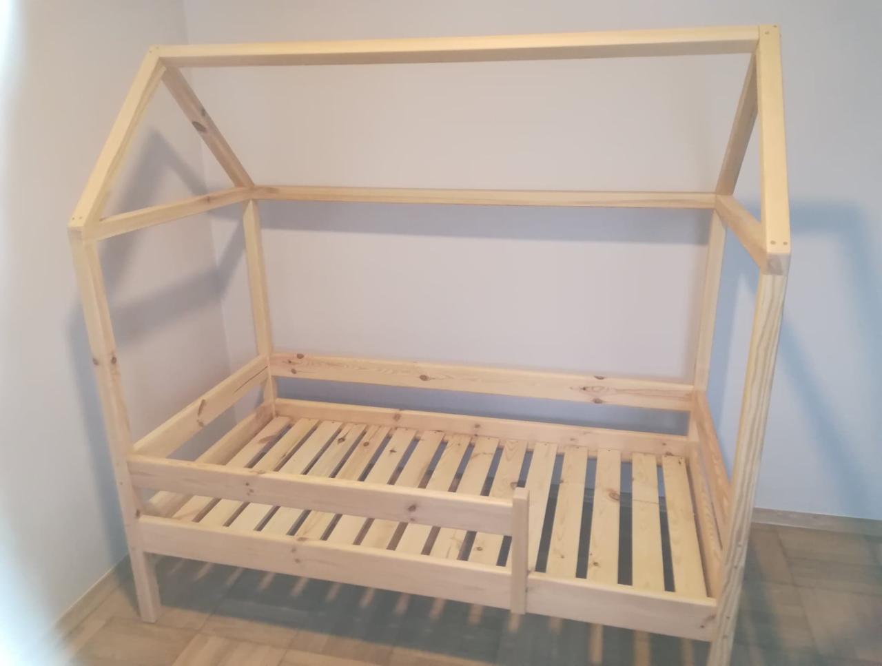 Surowa drewniana rama łóżka dziecięcego w kształcie domku, z listewkowym stelażem, widoczna na tle jasnej ściany i podłogi.