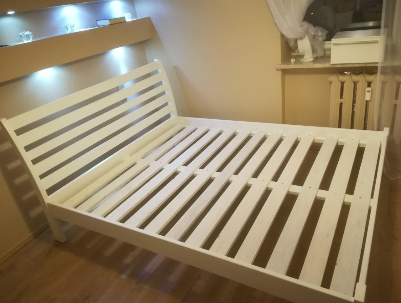 Biały, drewniany stelaż łóżka z listwami, widok z boku, w tle okno z firanką i grzejnik, oświetlenie górne.