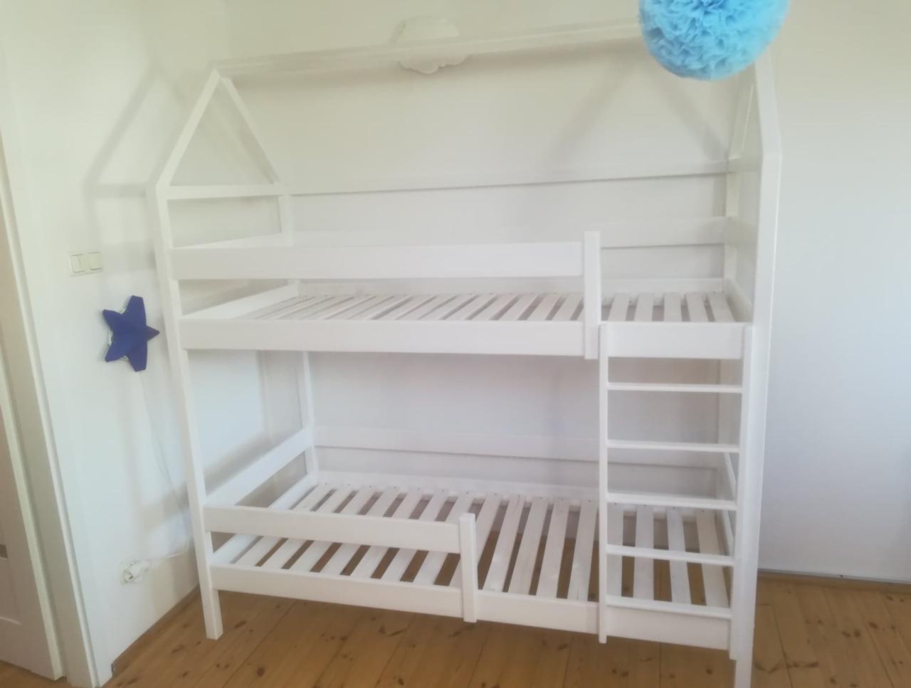 Białe, drewniane łóżko piętrowe w kształcie domku z drabinką, ozdobione niebieską gwiazdą na ścianie i pomponem u góry, na drewnianej podłodze.