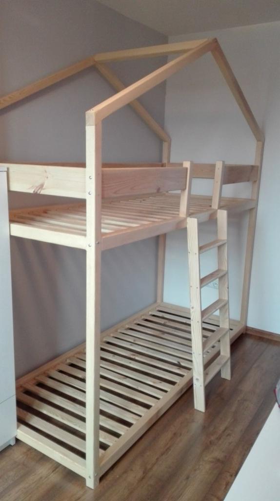 Drewniane łóżko piętrowe w kształcie domku, z drabinką, wykonane z jasnego drewna, ustawione na panelach podłogowych w pokoju z szarymi ścianami.