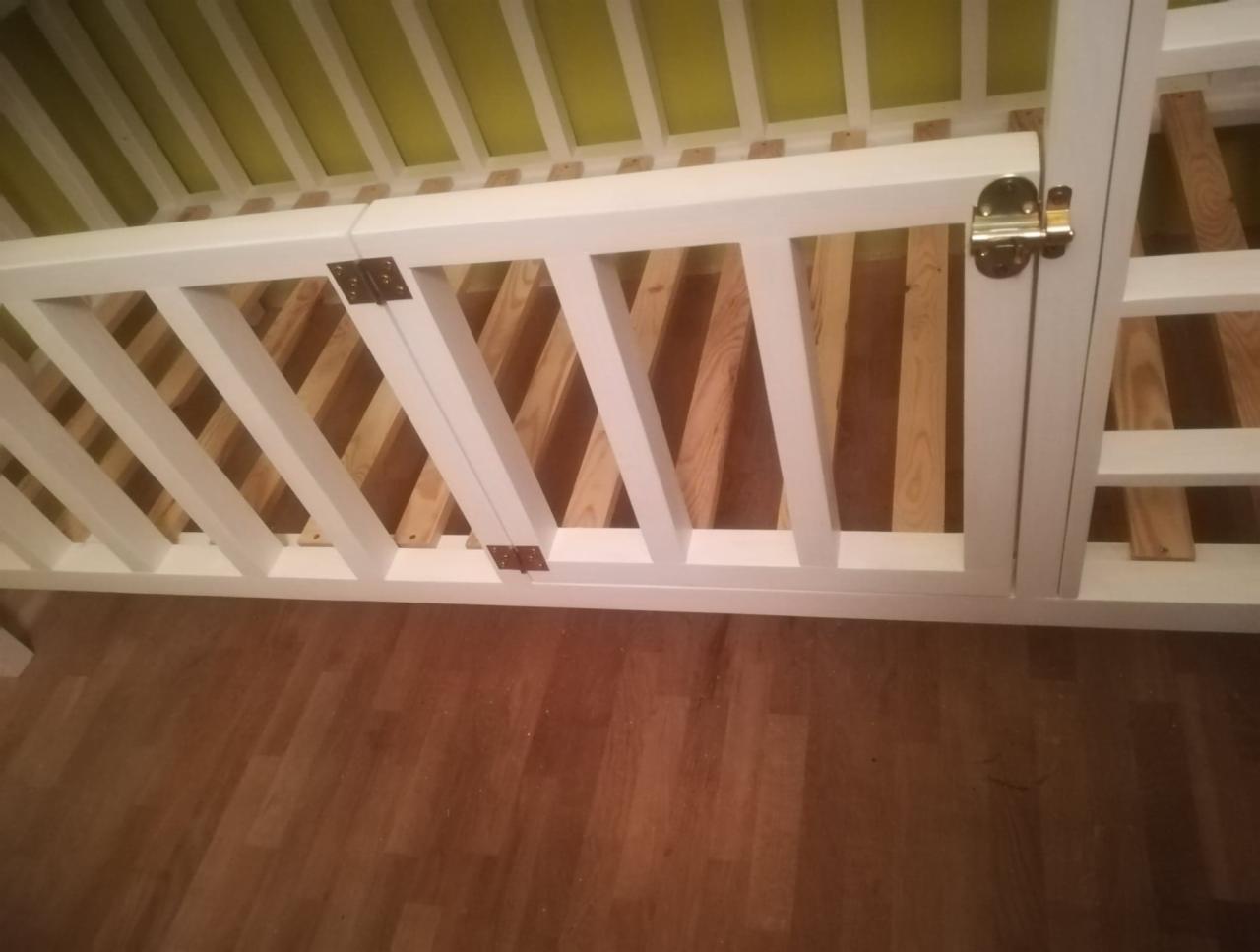 Biała konstrukcja drewniana z widocznymi zawiasami i złotym zamkiem, prawdopodobnie element antresoli lub mebla na wymiar, na tle drewnianej podłogi.