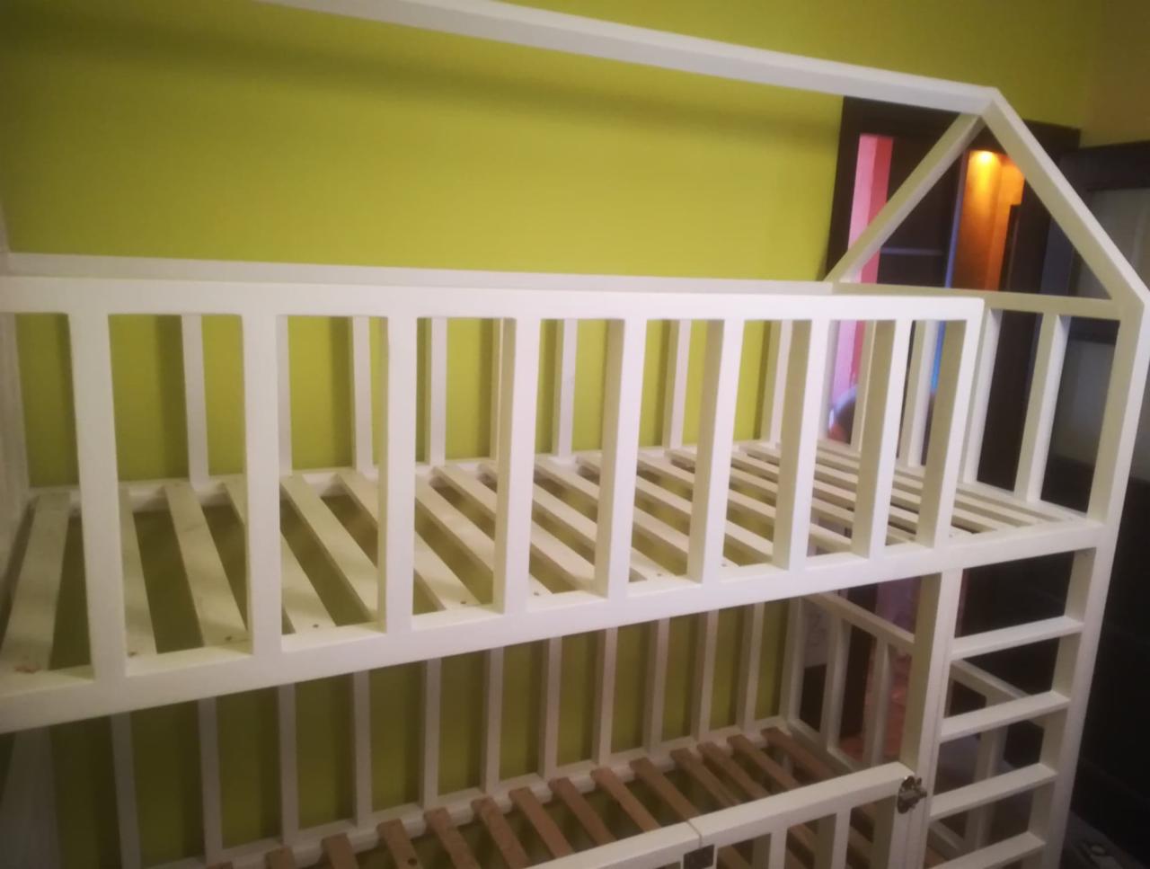 Białe, dwupoziomowe łóżko w kształcie domku z drabinką, na tle oliwkowozielonej ściany, widoczne drewniane listwy stelaża.