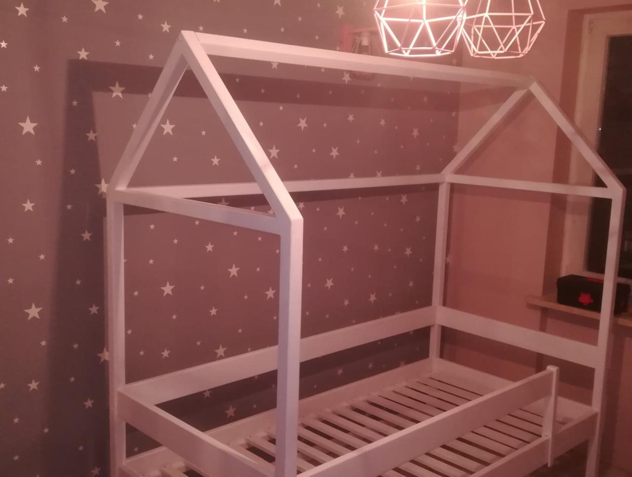 Białe, drewniane łóżko dziecięce w kształcie domku, z drabinką i stelażem, na tle ściany w gwiazdki i geometrycznych lamp