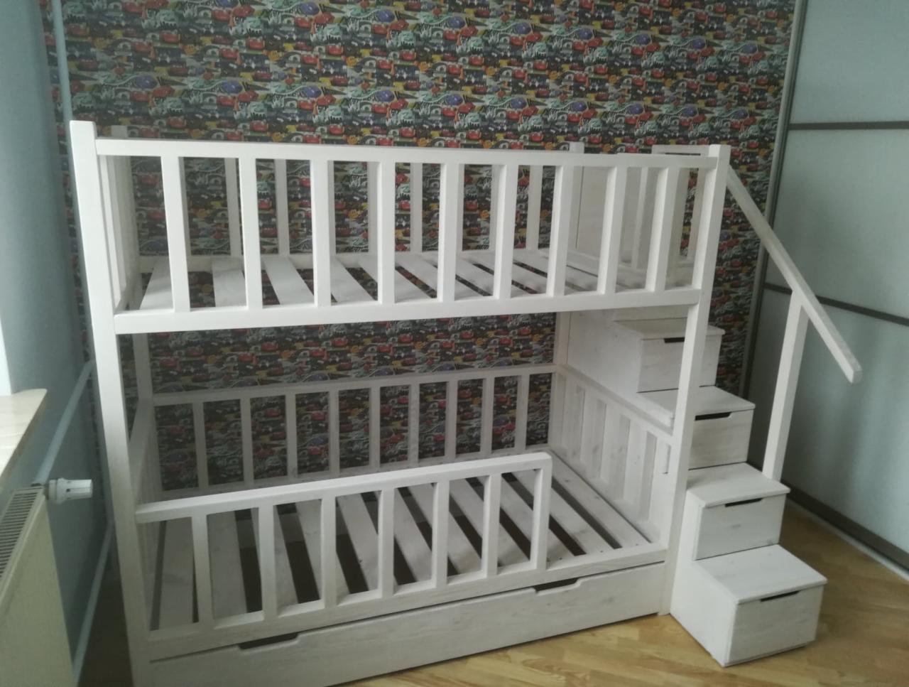 Białe łóżko piętrowe z drabinką w formie schodków z szufladami, na tle tapety w dziecięce wzory.