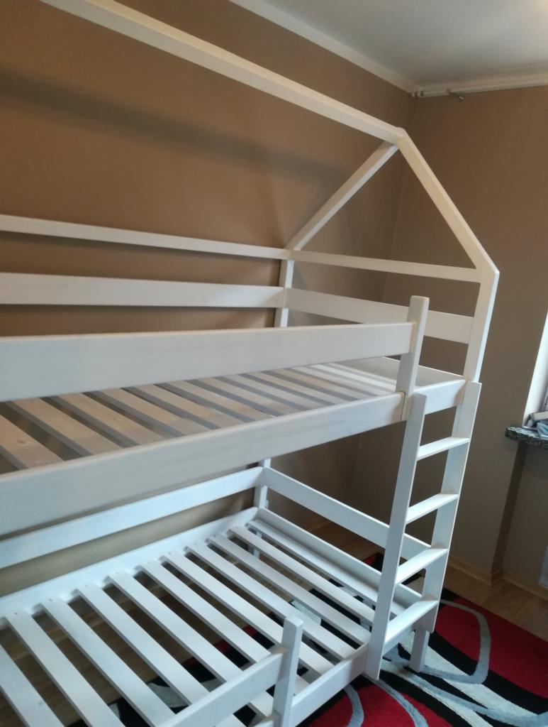 Białe łóżko piętrowe z drewnianą konstrukcją w kształcie domku, bez materacy, na czerwono-czarnym dywanie.