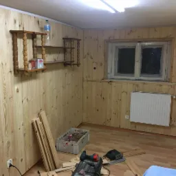 Wnętrze pokoju wykończonego boazerią w trakcie prac w Poznaniu, widoczne drewniane półki na ścianie, okno z grzejnikiem oraz narzędzia stolarskie na podłodze.