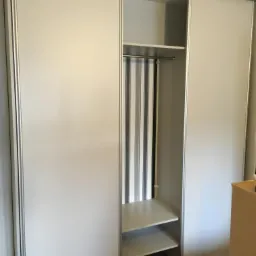 Wnętrze szafy przesuwnej z białymi drzwiami i aluminiowymi ramami, widoczne półki i drążek z zasłoną w pionowe paski.