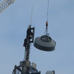 Montaż elementu anteny telekomunikacyjnej na wysokości; pracownicy w kaskach wspinają się po drabince, w tle błękitne niebo i fragment dźwigu.