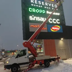 Pracownik na podnośniku koszowym, na tle ściany galerii handlowej z neonami znanych marek, ośnieżony plac.