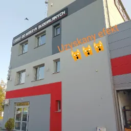 Budynek firmy z szaro-czerwoną elewacją, z napisem 'Części do maszyn budowlanych' i logo na dachu, widok z zewnątrz.