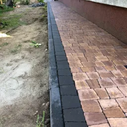 Ułożona ścieżka z kostki brukowej w odcieniach brązu i czerni, obok budynku z widocznym fragmentem ogrodu i sprzętu budowlanego w tle.