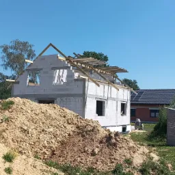 Budowa domu jednorodzinnego w trakcie wznoszenia, widoczna konstrukcja z pustaków i drewniana więźba dachowa, z przodu hałda piasku.