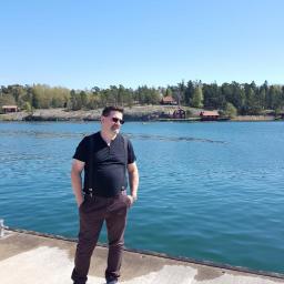 USŁUGI OGÓLNOBUDOWLANE Krzysztof Ptaś - Mężczyzna w szelkach i okularach przeciwsłonecznych stoi na betonowym nabrzeżu z widokiem na wodę i zabudowania w oddali w słoneczny dzień.