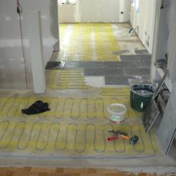 USŁUGI OGÓLNOBUDOWLANE Krzysztof Ptaś - Montaż ogrzewania podłogowego: żółta mata grzewcza z niebieskimi przewodami rozłożona na podłodze, częściowo przykryta szarymi płytkami, obok wiadro z zaprawą i narzędzia glazurnicze.