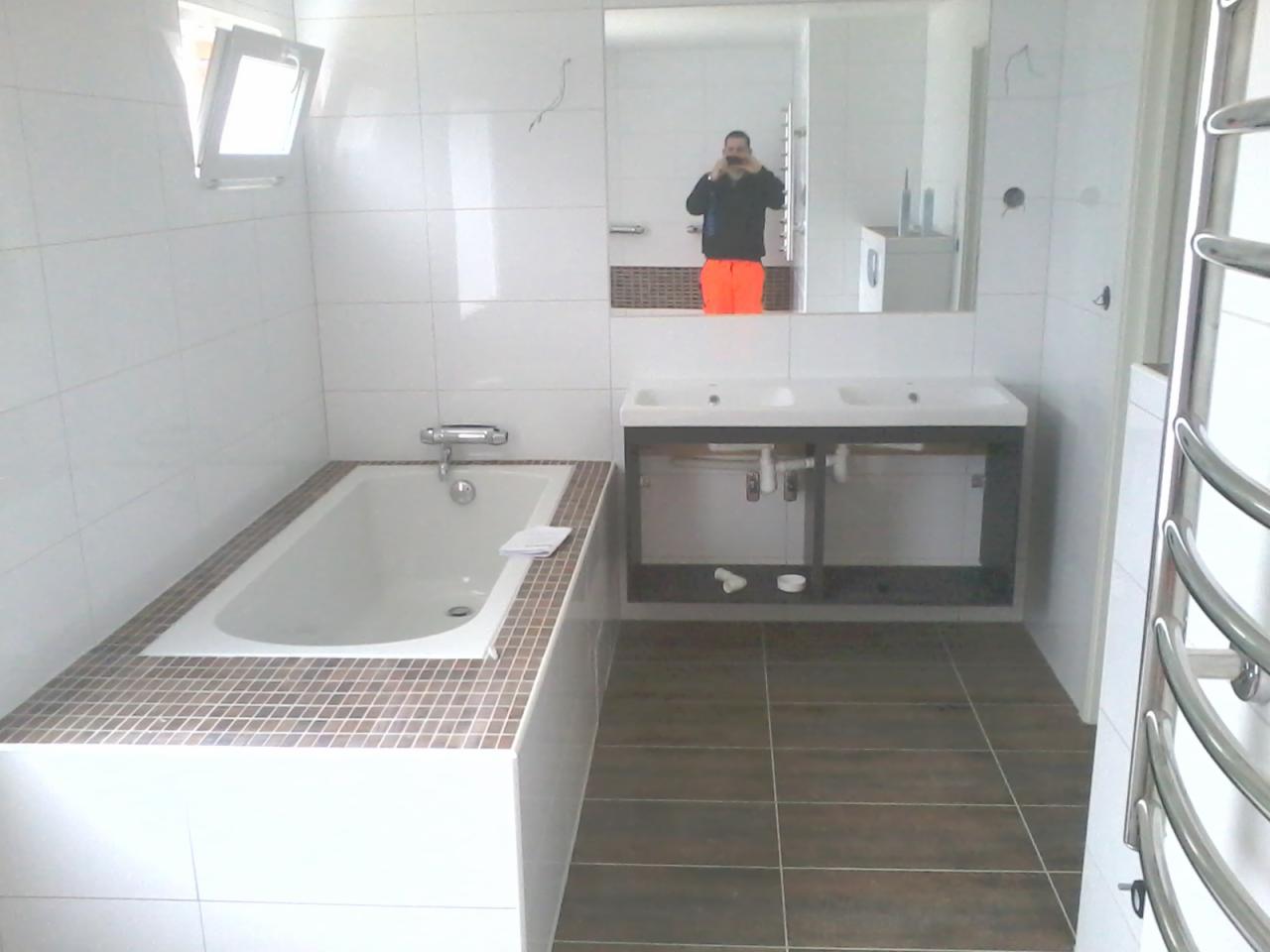 Wykończona łazienka z białą wanną obudowaną mozaiką, podwójną umywalką z szafką, brązowymi płytkami podłogowymi i grzejnikiem drabinkowym, widoczne odbicie osoby robiącej zdjęcie w lustrze.