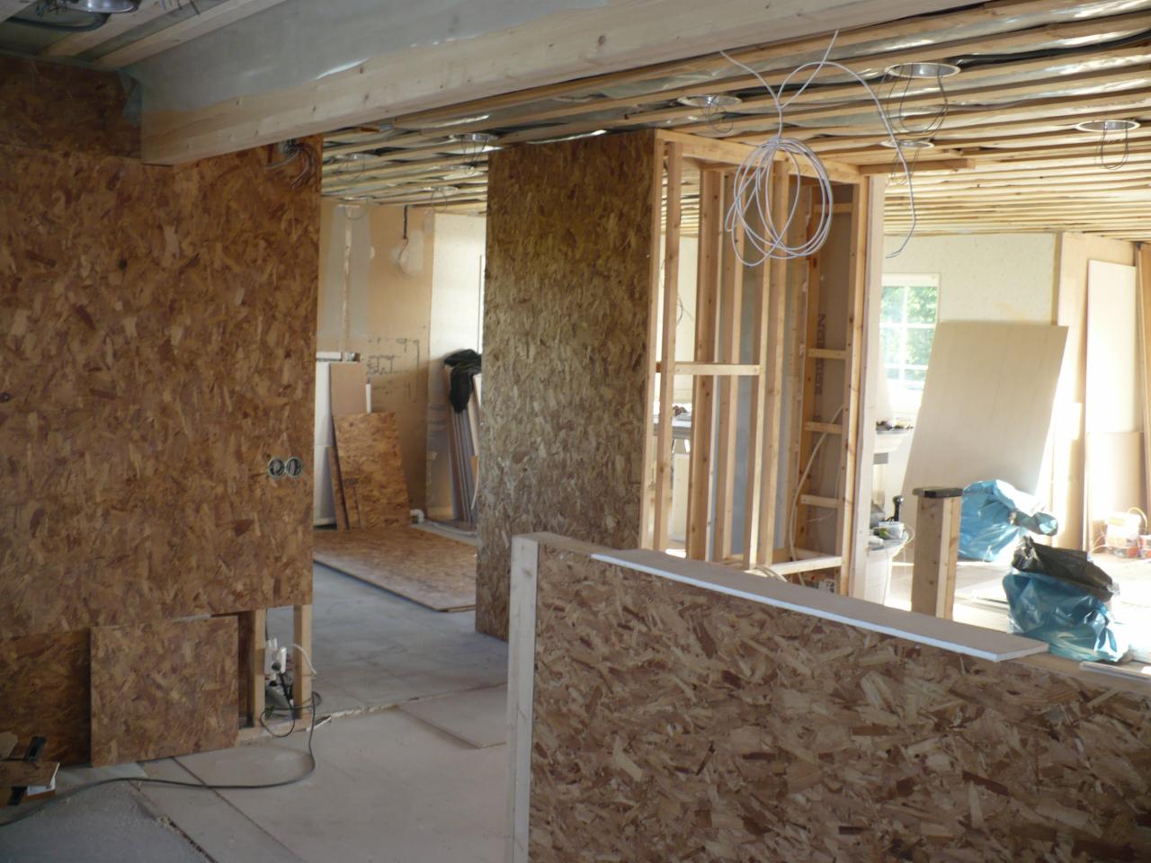 Wnętrze w trakcie remontu: widoczne drewniane konstrukcje ścian działowych, płyty OSB, przewody elektryczne wystające z sufitu, oraz surowy beton na podłodze.