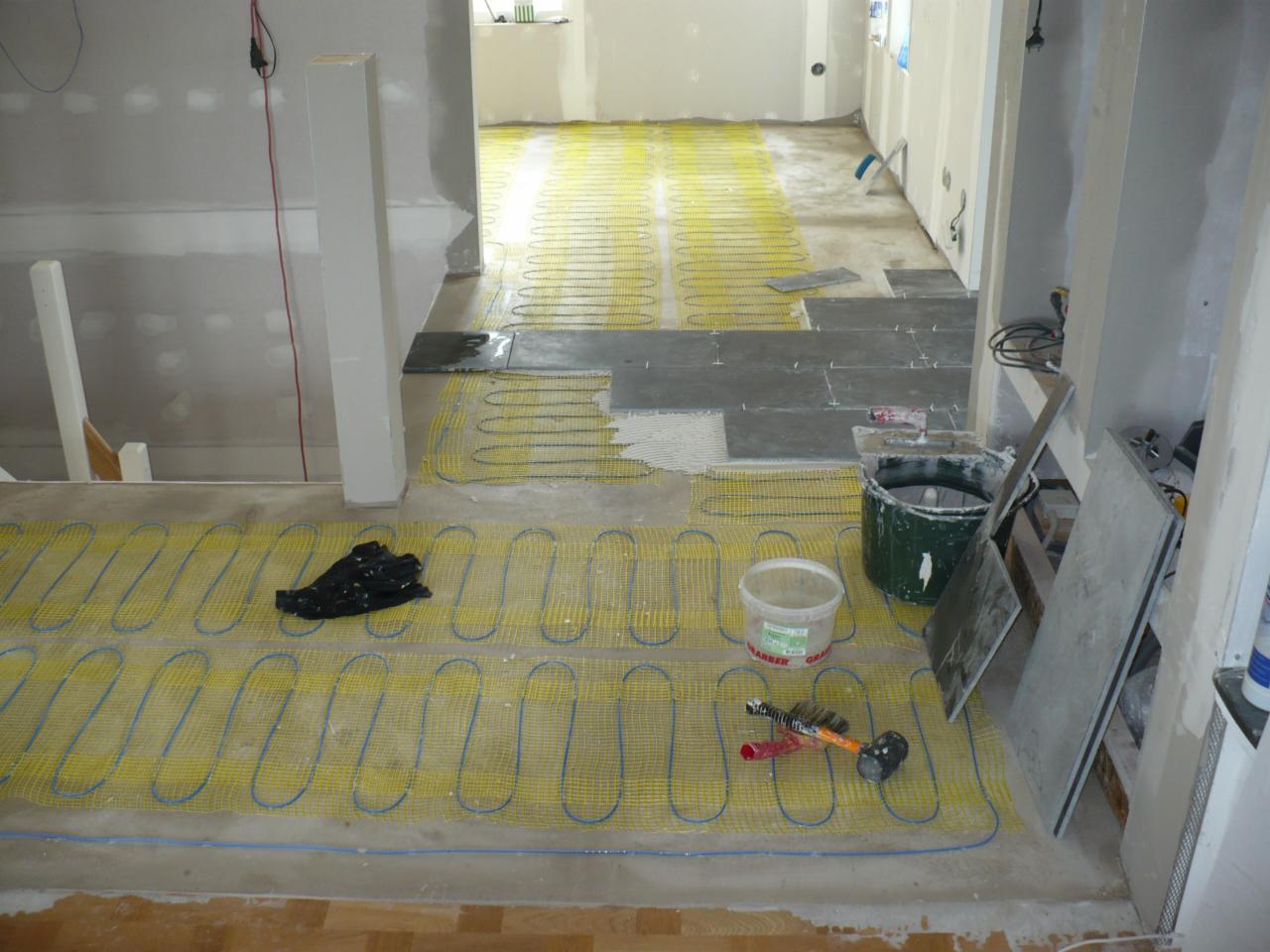 Montaż ogrzewania podłogowego: żółta mata grzewcza z niebieskimi przewodami rozłożona na podłodze, częściowo przykryta szarymi płytkami, obok wiadro z zaprawą i narzędzia glazurnicze.