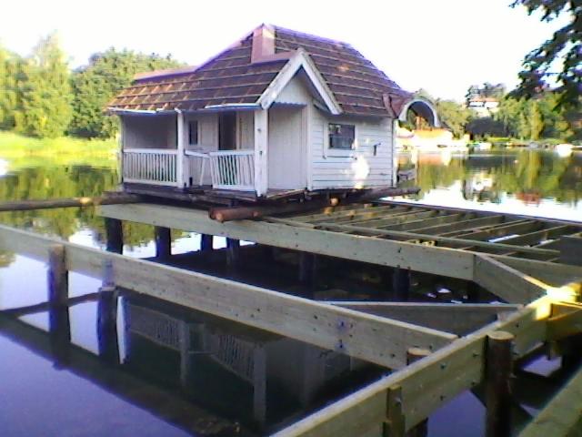 Konstrukcja drewnianego pomostu na wodzie z małym domkiem na palach w tle, widoczne odbicie w wodzie, drewniane elementy konstrukcyjne.