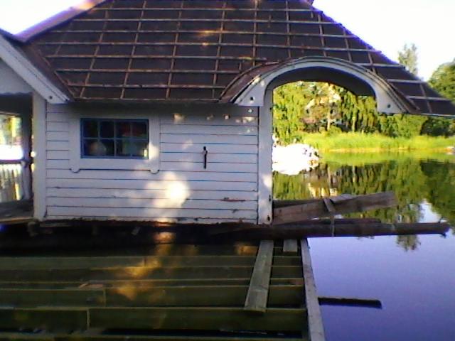 Niedokończona konstrukcja drewniana nad wodą, z widocznym zadaszeniem i elementami konstrukcyjnymi, w tle zieleń i odbicie w wodzie.