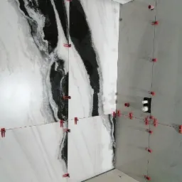 Świeżo ułożone płytki imitujące marmur na ścianie łazienki, z widocznymi plastikowymi krzyżykami dystansowymi i poziomnicą leżącą na podłodze.