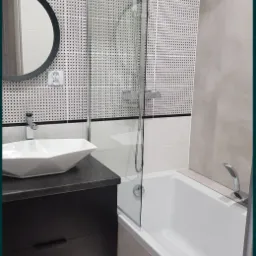 Nowoczesna łazienka z geometrycznym wzorem na ścianie, szklanym panelem prysznicowym, owalnym lustrem i podświetlaną szafką pod umywalką.