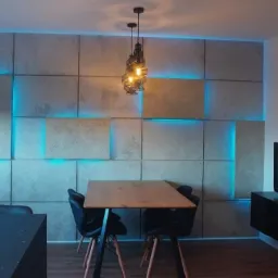 Ściana z paneli imitujących beton, podświetlona niebieskim światłem LED, drewniany stół z czarnymi krzesłami, wisząca lampa ze spiralnym kloszem, czarna szafka RTV z telewizorem, parkiet na podłodze.