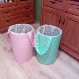 Dwa cylindryczne pojemniki z tkaniny w kolorach różowym i miętowym, ozdobione białymi gwiazdkami, z szarym wnętrzem w gwiazdki, stoją na podłodze obok drewnianych komód.
