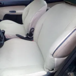 Jasne pokrowce z pikowaniem w romby na przednich fotelach samochodu, widoczne elementy deski rozdzielczej i fragment ściany z cegły w tle.