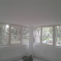 Jasne, nowo pomalowane wnętrze z dużymi oknami, widoczne grzejniki i pozostałości po remoncie na zewnątrz.