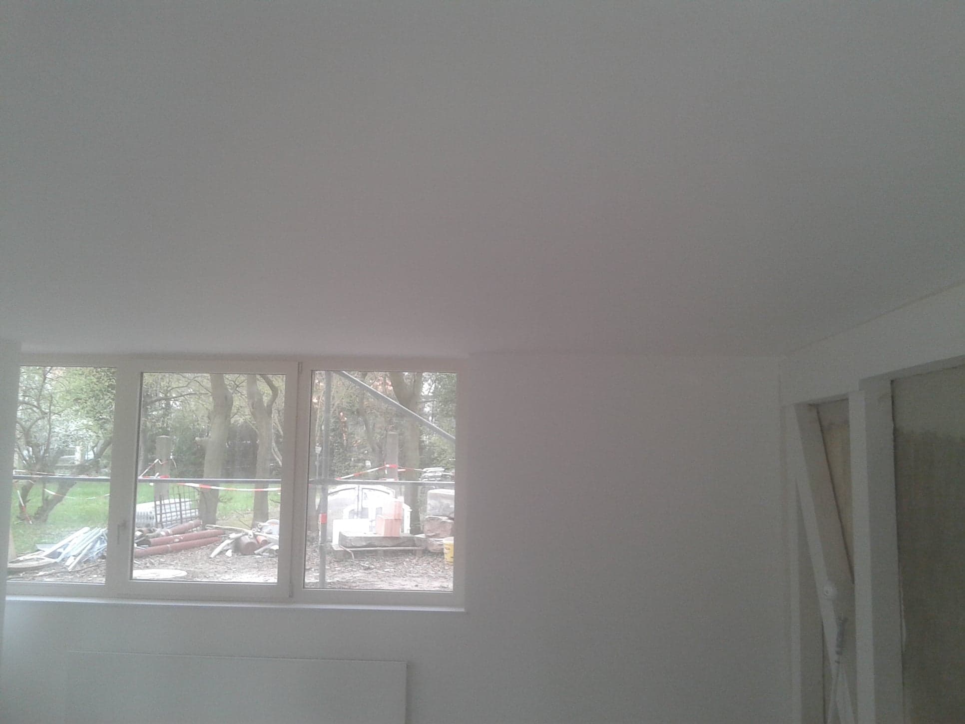 Wnętrze pokoju po pracach wykończeniowych: świeżo pomalowane ściany i sufit na biało, widok na zewnątrz przez okno z elementami budowlanymi na trawniku, widoczna ościeżnica drzwi.