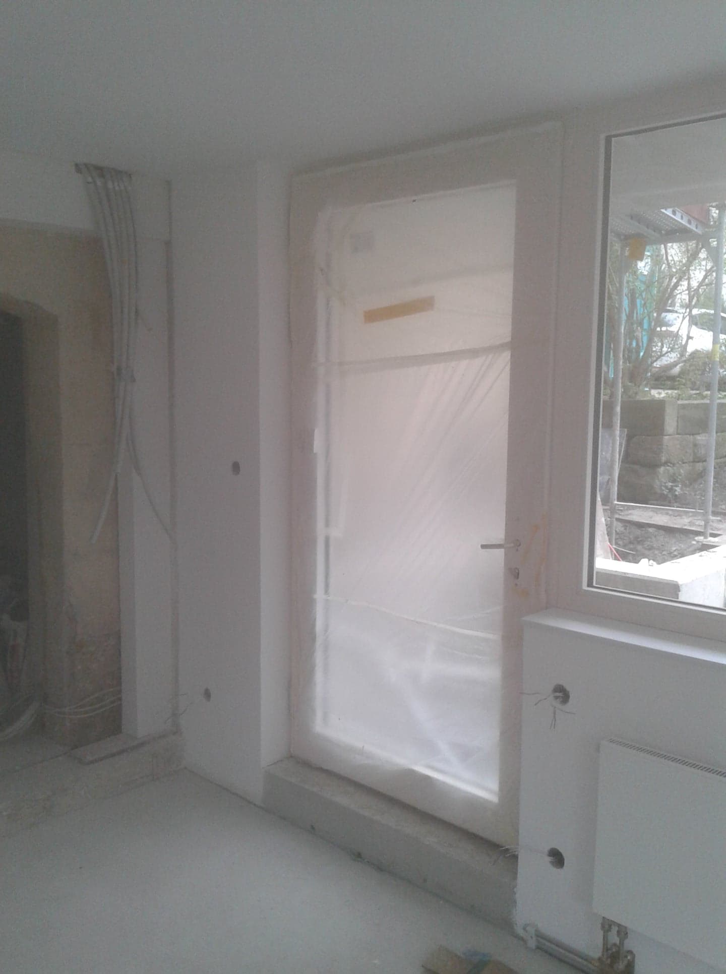 Wnętrze w trakcie remontu: zabezpieczone folią drzwi balkonowe, widoczne przewody elektryczne, surowa ściana i grzejnik, okno z widokiem na podwórze z rusztowaniem.