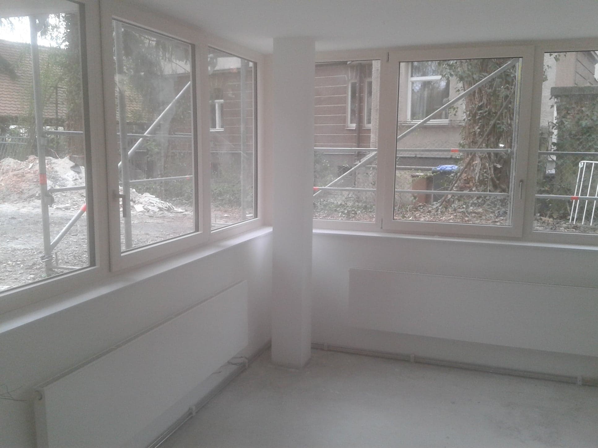 Wnętrze pokoju po remoncie, z świeżo pomalowanymi ścianami na biało, grzejnikami pod oknami i widokiem na rusztowania na zewnątrz.