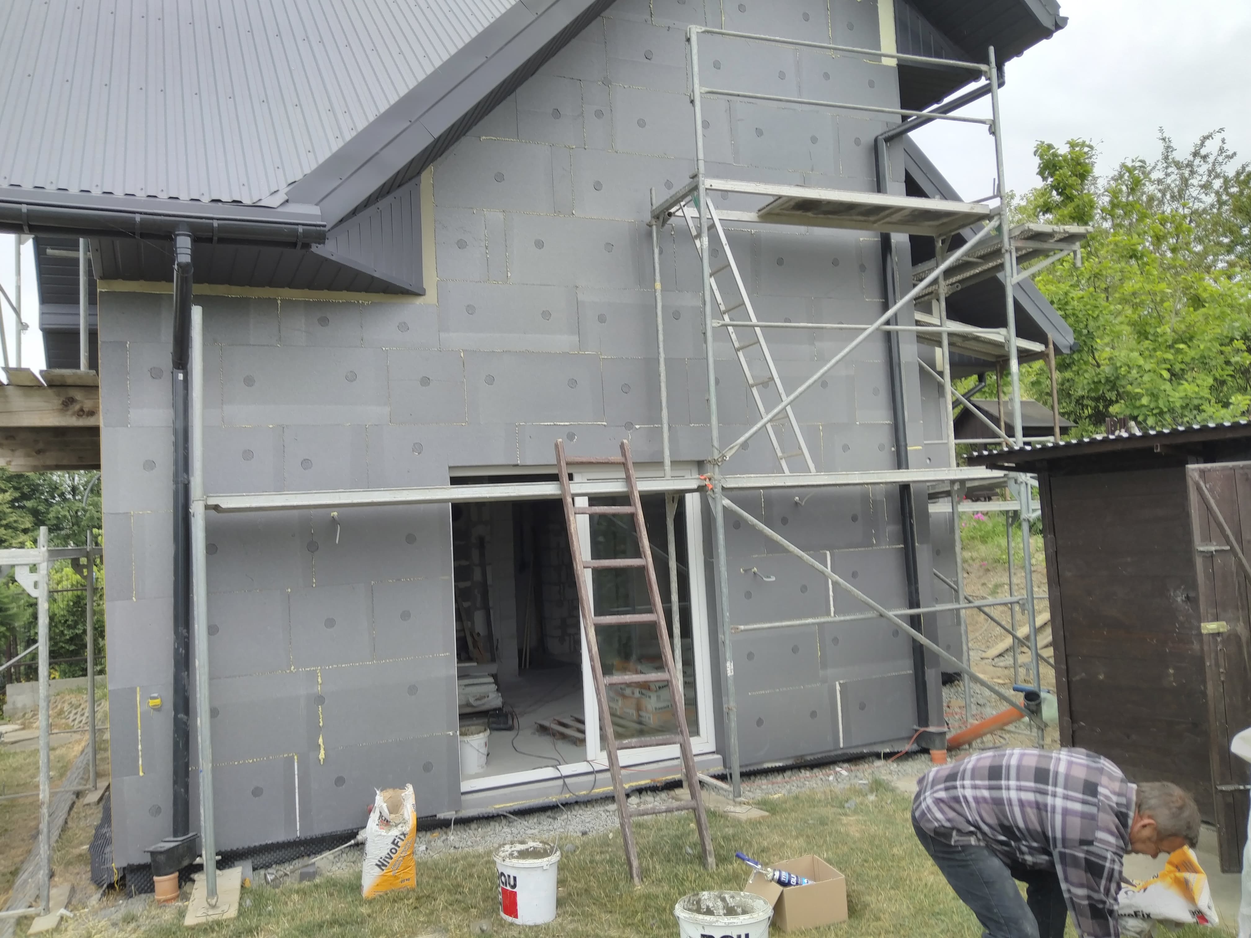 Dom w trakcie ocieplania ścian zewnętrznych, widoczne szare płyty izolacyjne, rusztowanie, drabina oparta o ścianę, worki z zaprawą i wiaderka z klejem, pracownik w koszuli w kratę.