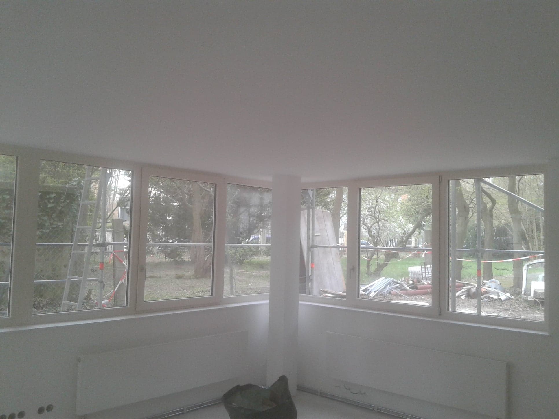 Jasne, nowo pomalowane wnętrze z dużymi oknami, widoczne grzejniki i pozostałości po remoncie na zewnątrz.