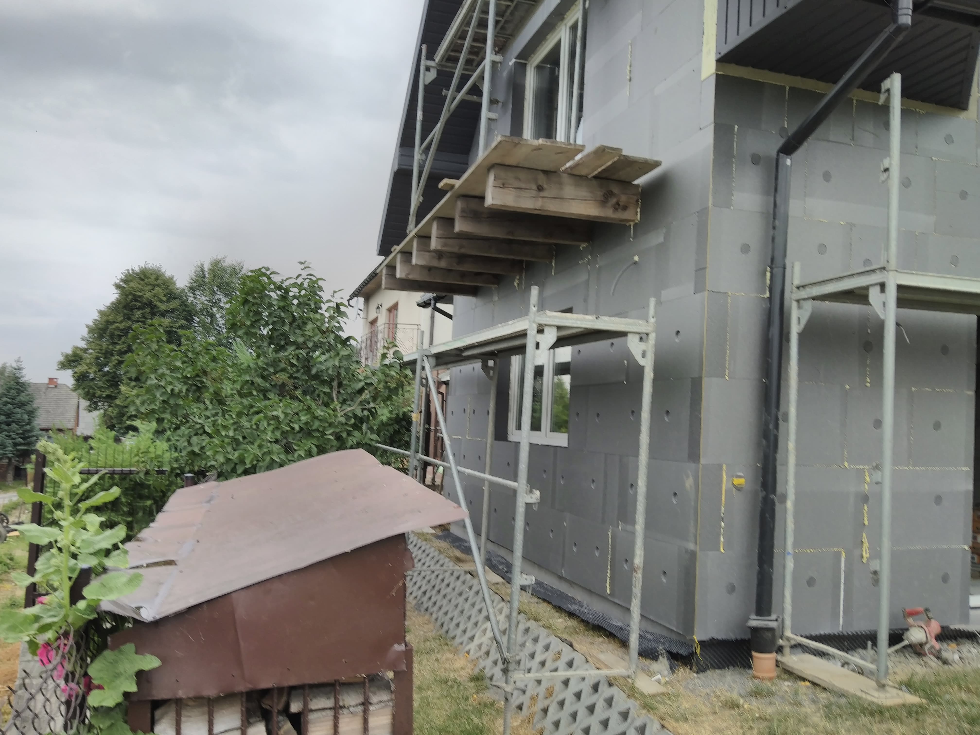 Dom w trakcie ocieplania ścian zewnętrznych styropianem, widoczne rusztowania i drewniane pomosty, częściowo zakończone prace, szare płyty izolacyjne z widocznymi otworami.