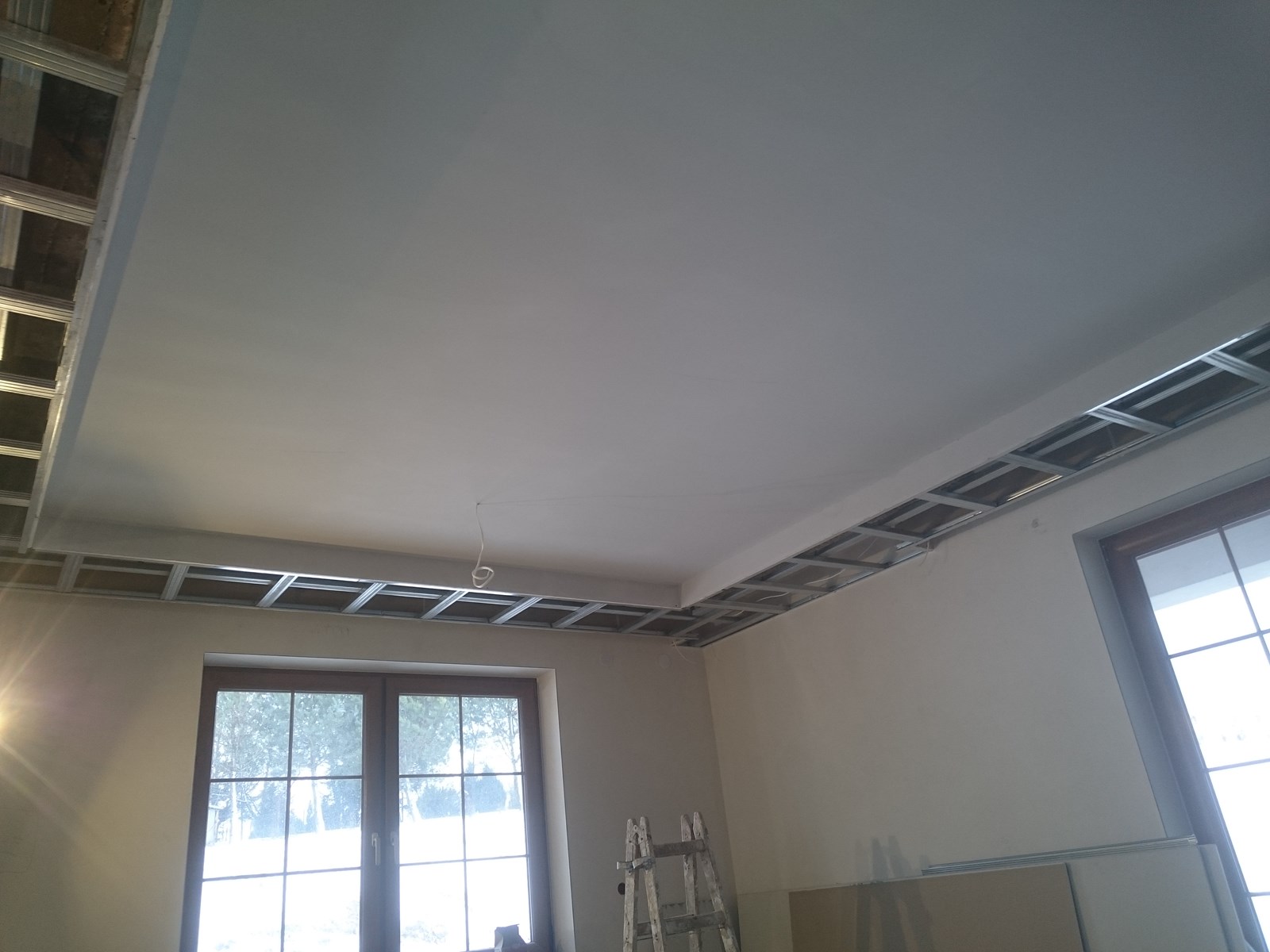 Sufit podwieszany w trakcie montażu, widoczna metalowa konstrukcja rusztowa, częściowo zakryta płytami gipsowo-kartonowymi, drabina i okna w tle.