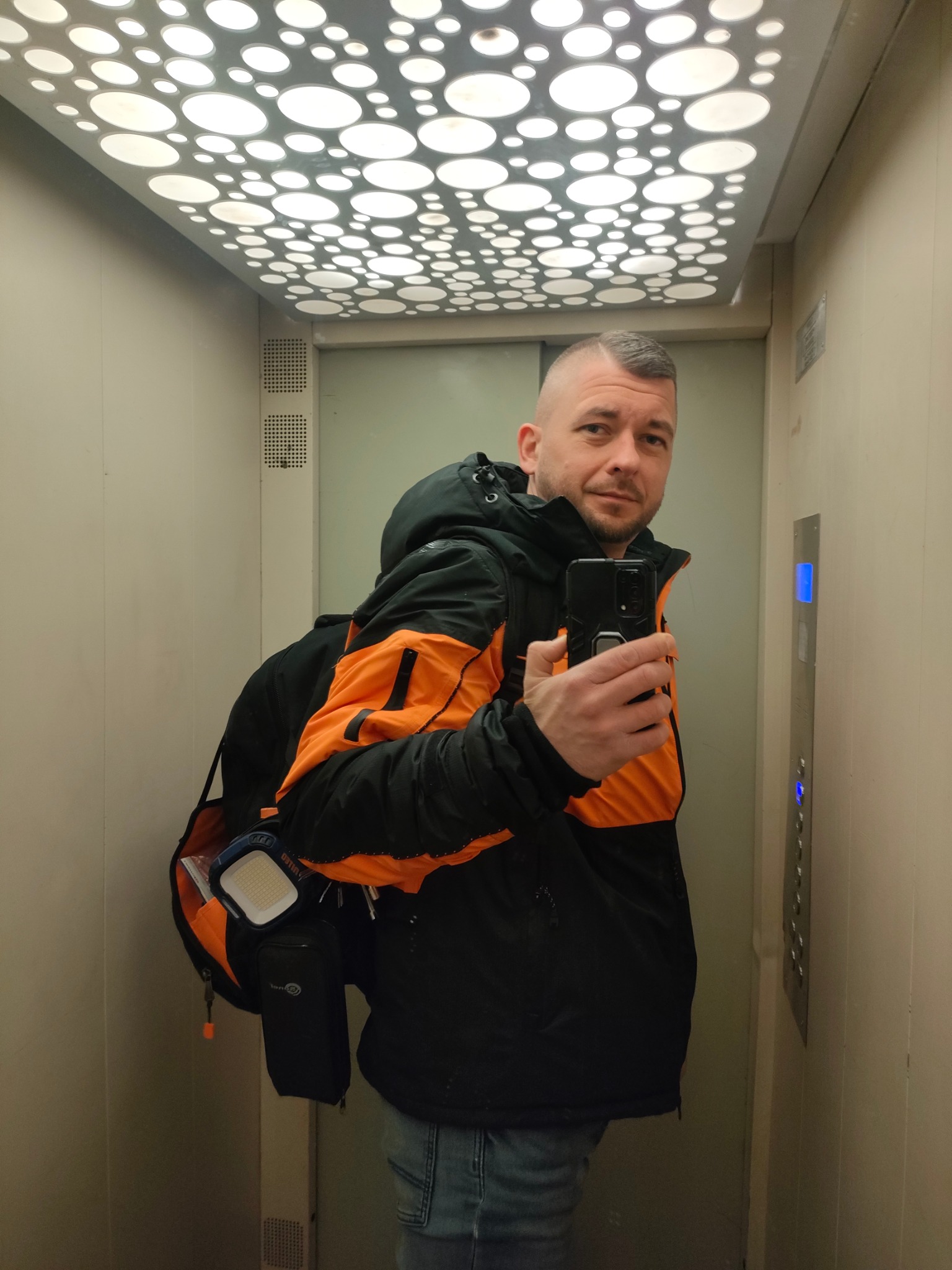 Mężczyzna w kurtce roboczej z torbą narzędziową na ramieniu robi selfie w windzie z dekoracyjnym oświetleniem sufitowym i przyciskami windy.
