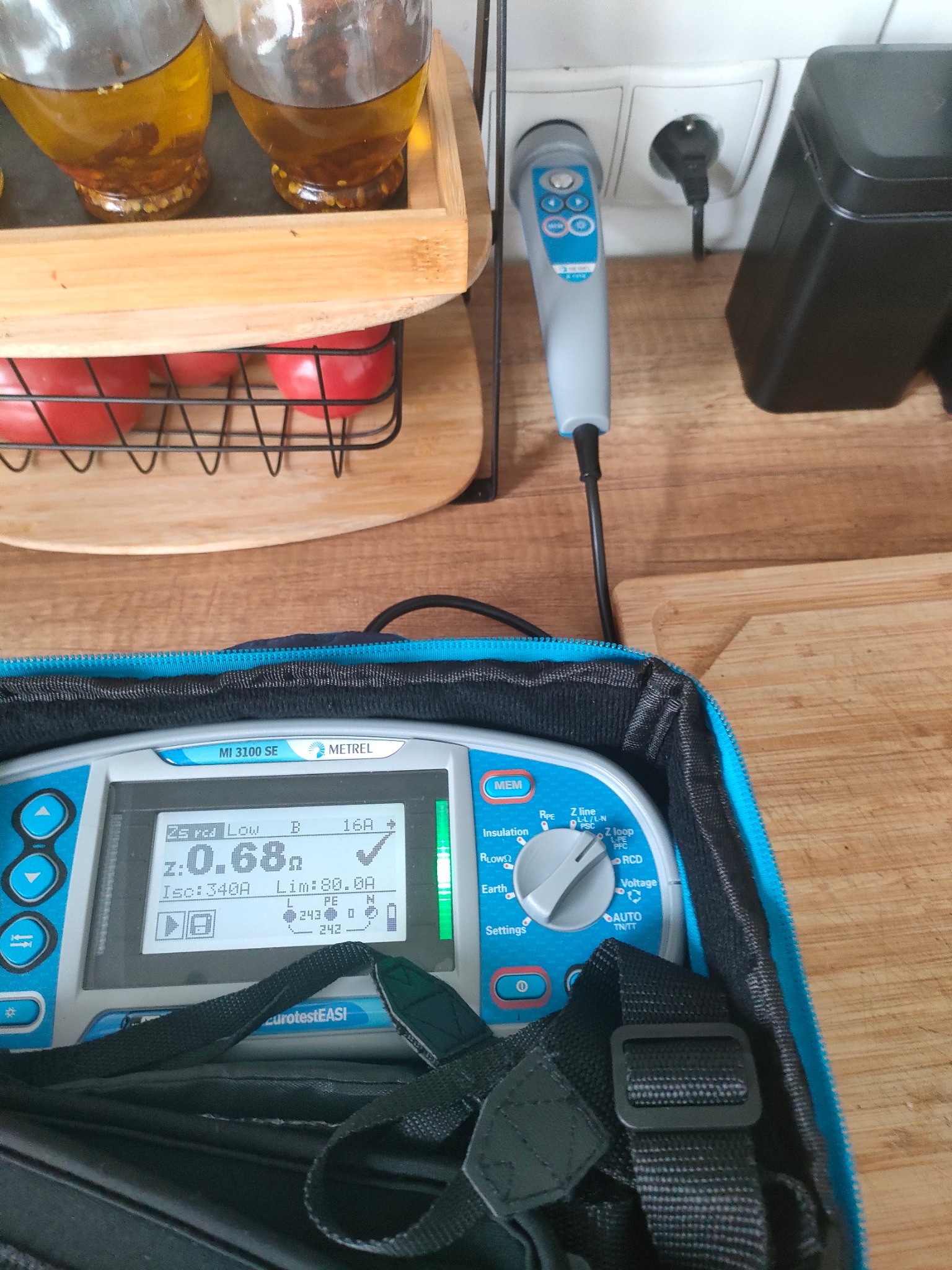 Miernik elektryczny Metrel MI 3100 SE w niebieskim futerale, sonda w gniazdku elektrycznym, pomiar impedancji pętli zwarcia widoczny na ekranie, w tle przyprawy w oleju i pomidory.