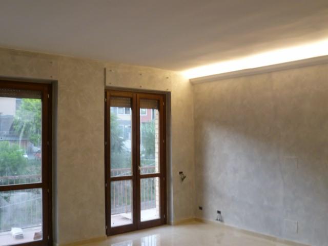 Wnętrze pokoju z wykończonymi ścianami w odcieniach szarości i beżu, imitującymi efekt dekoracyjny. Widoczne drewniane ramy okienne i drzwi balkonowe, listwa oświetleniowa pod sufitem oraz jasna...