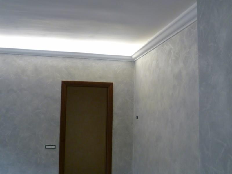 Wnętrze pokoju z dekoracyjną, szarą ścianą o efekcie marmuru, białym sztukateryjnym gzymsem oświetlonym od góry, brązowymi drzwiami i minimalistycznym gniazdkiem elektrycznym.